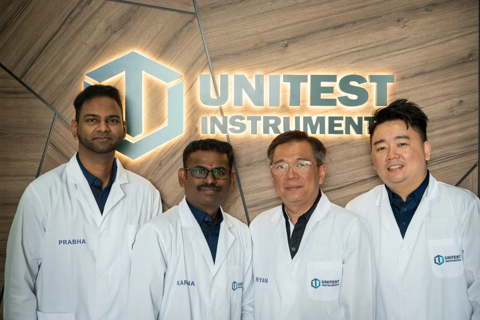 The Unitest Instruments calibration team in white lab coats at our Singapore office, beneath the illuminated Unitest Instruments logo wall